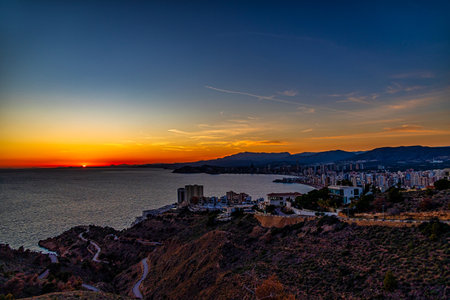 A stunning sunset casts a warm glow over a coastal city, with the sun reflecting beautifully on the calm seaの写真素材