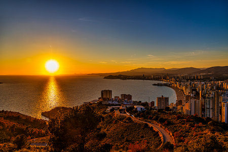 A stunning sunset casts a warm glow over a coastal city, with the sun reflecting beautifully on the calm seaの写真素材