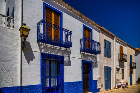 Whitewashed buildings with vibrant blue accents and wrought iron balconies line a sunlit streetの写真素材