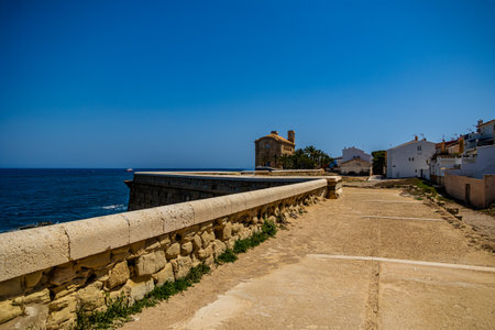 A stone path with a sturdy wall overlooks the ocean, leading toward a small coastal village with white buildingsの写真素材
