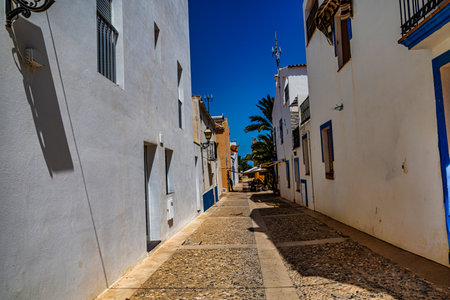 A narrow cobblestone alleyway is lined with simple, light-colored buildings under a clear blue sky.の写真素材
