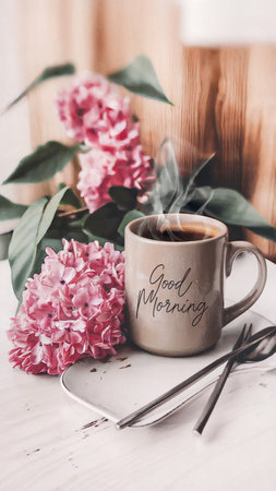 A steaming mug with Good Morning written on it sits on a white surface beside pink hydrangea flowers.の素材