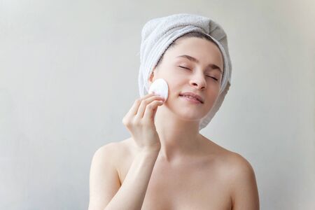 Beauty portrait of a smiling brunette woman in towel on head with soft healthy skin removing make up with cotton pad isolated over white background. Skincare cleansing spa relax conceptの写真素材