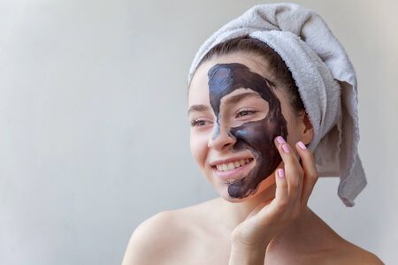 Beauty portrait of a smiling brunette woman in a towel on the head applying black nourishing mask on face on white background isolated. Skincare cleansing spa relax cosmetics conceptの写真素材