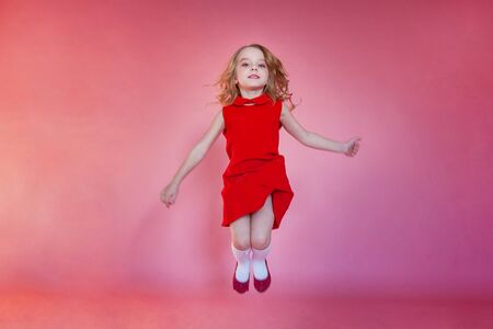 Little cute sweet smiling girl in red dress jumping on pink colourful pastel trendy modern fashion...