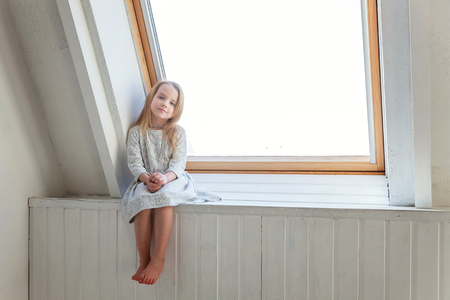 Little cute sweet smiling girl in white dress sitting on the window sill in bright light living room at home and thinkingの写真素材