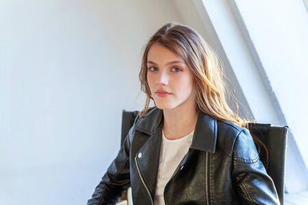 Young teenage girl in leather jacket sitting on modern chair posing in bright light room against...