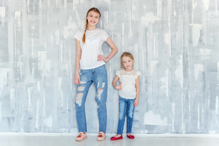 Two happy kids standing against grey textured wall background and embracing. Adorable pretty little girl hugging tight cute teenage girl, showing her love and care. Sisters having fun at homeの写真素材