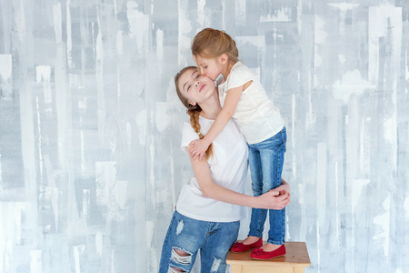 Two happy kids standing against grey textured wall background and embracing. Adorable pretty little girl hugging tight cute teenage girl, showing her love and care. Sisters having fun at homeの写真素材