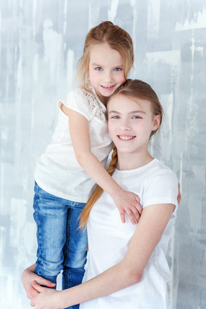 Two happy kids standing against grey textured wall background and embracing. Adorable pretty little girl hugging tight cute teenage girl, showing her love and care. Sisters having fun at homeの写真素材