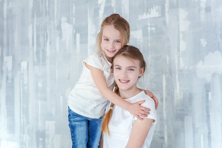 Two happy kids standing against grey textured wall background and embracing. Adorable pretty little girl hugging tight cute teenage girl, showing her love and care. Sisters having fun at homeの写真素材