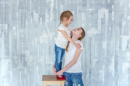 Two happy kids standing against grey textured wall background and embracing. Adorable pretty little girl hugging tight cute teenage girl, showing her love and care. Sisters having fun at homeの写真素材