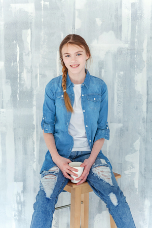 Young happy cute teenage girl in jeans, denim jacket and white T-shirt sitting on chair against grey textured wall background, holding paper cup and drinking coffee. Casual daily lifestyleの写真素材
