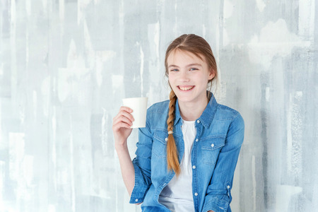Young happy cute teenage girl in jeans, denim jacket and white T-shirt sitting on chair against grey textured wall background, holding paper cup and drinking coffee. Casual daily lifestyleの写真素材