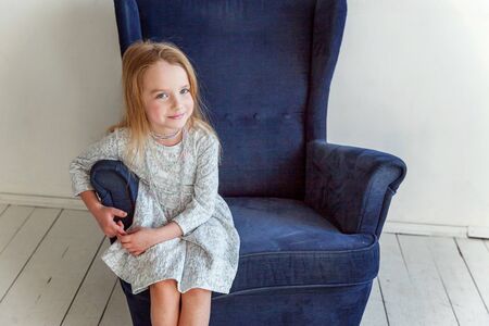 Sweet little girl in grey dress at home sitting on modern cozy blue chair relaxing in white living roomの写真素材