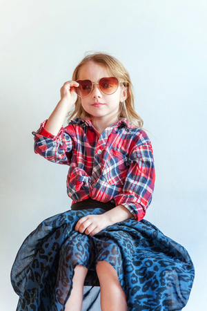 Sweet happy adorable fashion young little girl dressed like hipster hippie gipsy cowboy in sunglasses sitting on modern chair posing in white wall background in light bright room. Lifestyle people trendy conceptの写真素材