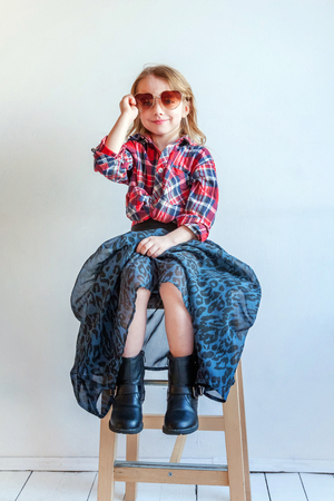 Sweet happy adorable fashion young little girl dressed like hipster hippie gipsy cowboy in sunglasses sitting on modern chair posing in white wall background in light bright room. Lifestyle people trendy conceptの写真素材
