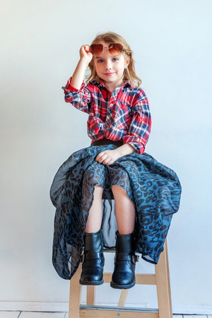 Sweet happy adorable fashion young little girl dressed like hipster hippie gipsy cowboy in sunglasses sitting on modern chair posing in white wall background in light bright room. Lifestyle people trendy conceptの写真素材