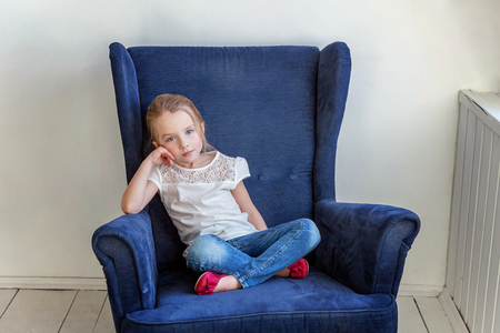 Sweet little girl in jeans and white T-shirt at home sitting on modern cozy blue chair relaxing in white living roomの写真素材