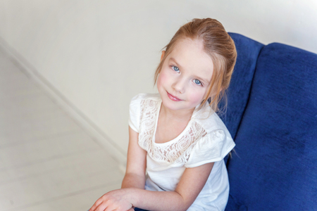 Sweet little girl in jeans and white T-shirt at home sitting on modern cozy blue chair relaxing in white living roomの写真素材