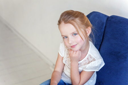 Sweet little girl in jeans and white T-shirt at home sitting on modern cozy blue chair relaxing in white living roomの写真素材