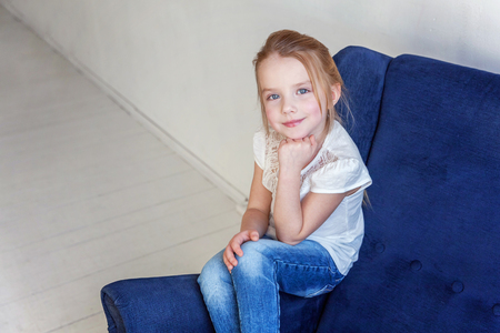 Sweet little girl in jeans and white T-shirt at home sitting on modern cozy blue chair relaxing in white living roomの写真素材