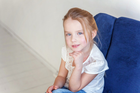 Sweet little girl in jeans and white T-shirt at home sitting on modern cozy blue chair relaxing in white living roomの写真素材