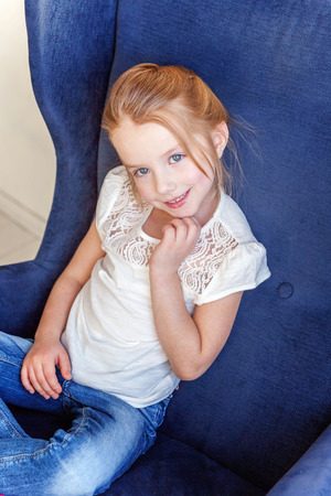 Sweet little girl in jeans and white T-shirt at home sitting on modern cozy blue chair relaxing in white living roomの写真素材
