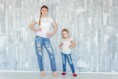 Two happy kids standing against grey textured wall background and embracing. Adorable pretty little girl hugging tight cute teenage girl, showing her love and care. Sisters having fun at homeの写真素材