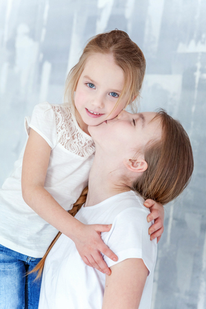 Two happy kids standing against grey textured wall background and embracing. Adorable pretty little girl hugging tight cute teenage girl, showing her love and care. Sisters having fun at homeの写真素材