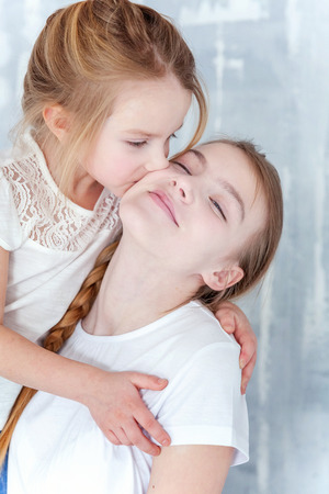 Two happy kids standing against grey textured wall background and embracing. Adorable pretty little girl hugging tight cute teenage girl, showing her love and care. Sisters having fun at homeの写真素材