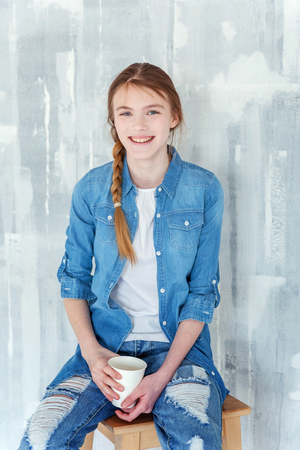 Young happy cute teenage girl in jeans, denim jacket and white T-shirt sitting on chair against grey textured wall background, holding paper cup and drinking coffee. Casual daily lifestyleの写真素材
