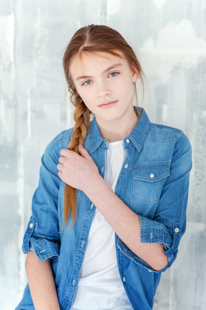 Young happy cute teenage girl in jeans, denim jacket and white T-shirt sitting on chair against grey textured wall background. Casual daily lifestyle. Schoolgirl student education and school conceptの写真素材
