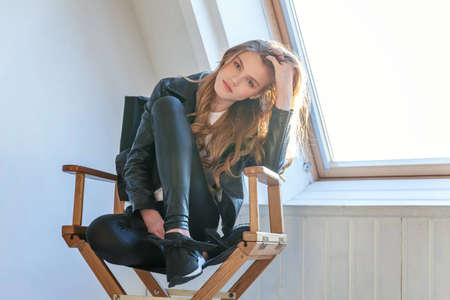 Young teenage girl in leather jacket sitting on modern chair posing in bright light room against...