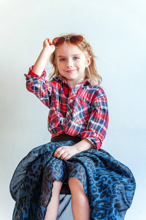 Sweet happy adorable fashion young little girl dressed like hipster hippie gipsy cowboy in sunglasses sitting on modern chair posing in white wall background in light bright room. Lifestyle people trendy conceptの写真素材
