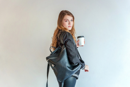 Young teenage girl in leather jacket against white wall, holding paper cup and drinking coffee. Casual daily lifestyle. Pretty sexy fashion sensual woman dressed in hipster rock style outfitの写真素材