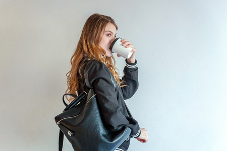 Young teenage girl in leather jacket against white wall, holding paper cup and drinking coffee. Casual daily lifestyle. Pretty sexy fashion sensual woman dressed in hipster rock style outfitの写真素材