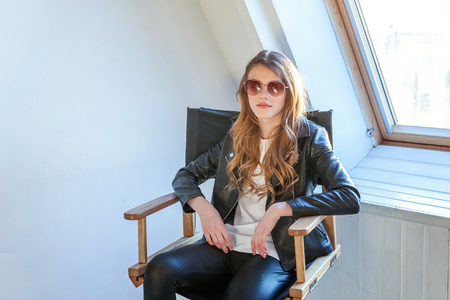 Young teenage girl in leather jacket sitting on modern chair posing in bright light room against white wall background window. Pretty sexy fashion sensual woman dressed in hipster rock style outfitの写真素材
