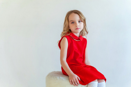 Sweet happy little girl in red dress sitting on chair against white wall background in bright roomの写真素材