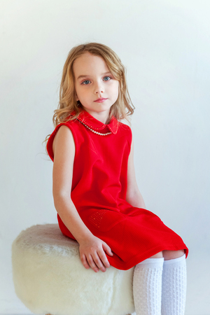 Sweet happy little girl in red dress sitting on chair against white wall background in bright roomの写真素材