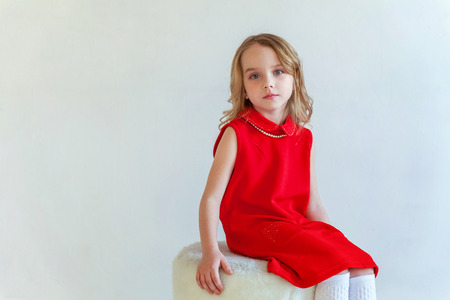Sweet happy little girl in red dress sitting on chair against white wall background in bright roomの写真素材