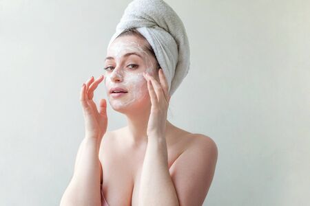 Beauty portrait of a smiling brunette woman in a towel on the head applying white nourishing mask or creme on face on white background isolated. Skincare cleansing spa relax conceptの写真素材