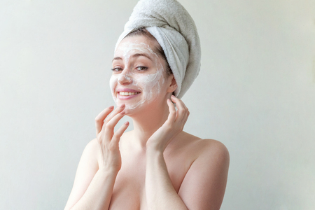 Beauty portrait of a smiling brunette woman in a towel on the head applying white nourishing mask or creme on face on white background isolated. Skincare cleansing spa relax conceptの写真素材
