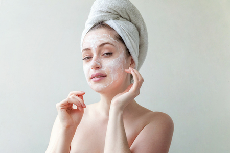 Beauty portrait of a smiling brunette woman in a towel on the head applying white nourishing mask or creme on face on white background isolated. Skincare cleansing spa relax conceptの写真素材