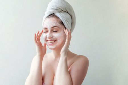 Beauty portrait of a smiling brunette woman in a towel on the head applying white nourishing mask or creme on face on white background isolated. Skincare cleansing spa relax conceptの写真素材