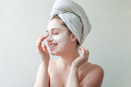 Beauty portrait of a smiling brunette woman in a towel on the head applying white nourishing mask or creme on face on white background isolated. Skincare cleansing spa relax conceptの写真素材