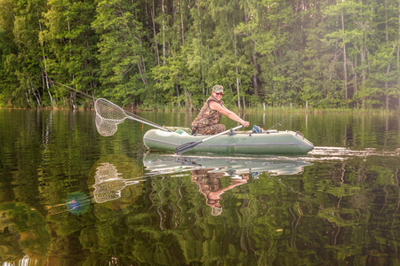 Fisherman with fishing rods is fishing in a rubber boat against background of beautiful nature and lake or river. Camping tourism relax trip active lifestyle adventure conceptの写真素材