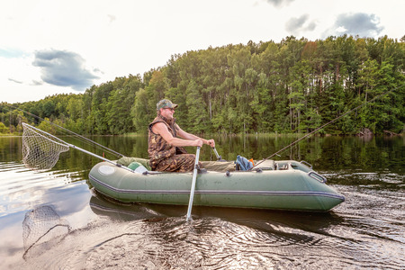 Fisherman with fishing rods is fishing in a rubber boat against background of beautiful nature and lake or river. Camping tourism relax trip active lifestyle adventure conceptの写真素材