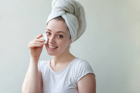 Beauty portrait of a smiling brunette woman with towel on head with soft healthy skin removing make up with cotton pad isolated over white background. Skincare cleansing spa relax conceptの写真素材