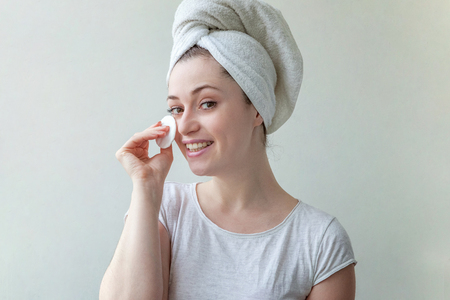 Beauty portrait of a smiling brunette woman with towel on head with soft healthy skin removing make up with cotton pad isolated over white background. Skincare cleansing spa relax conceptの写真素材